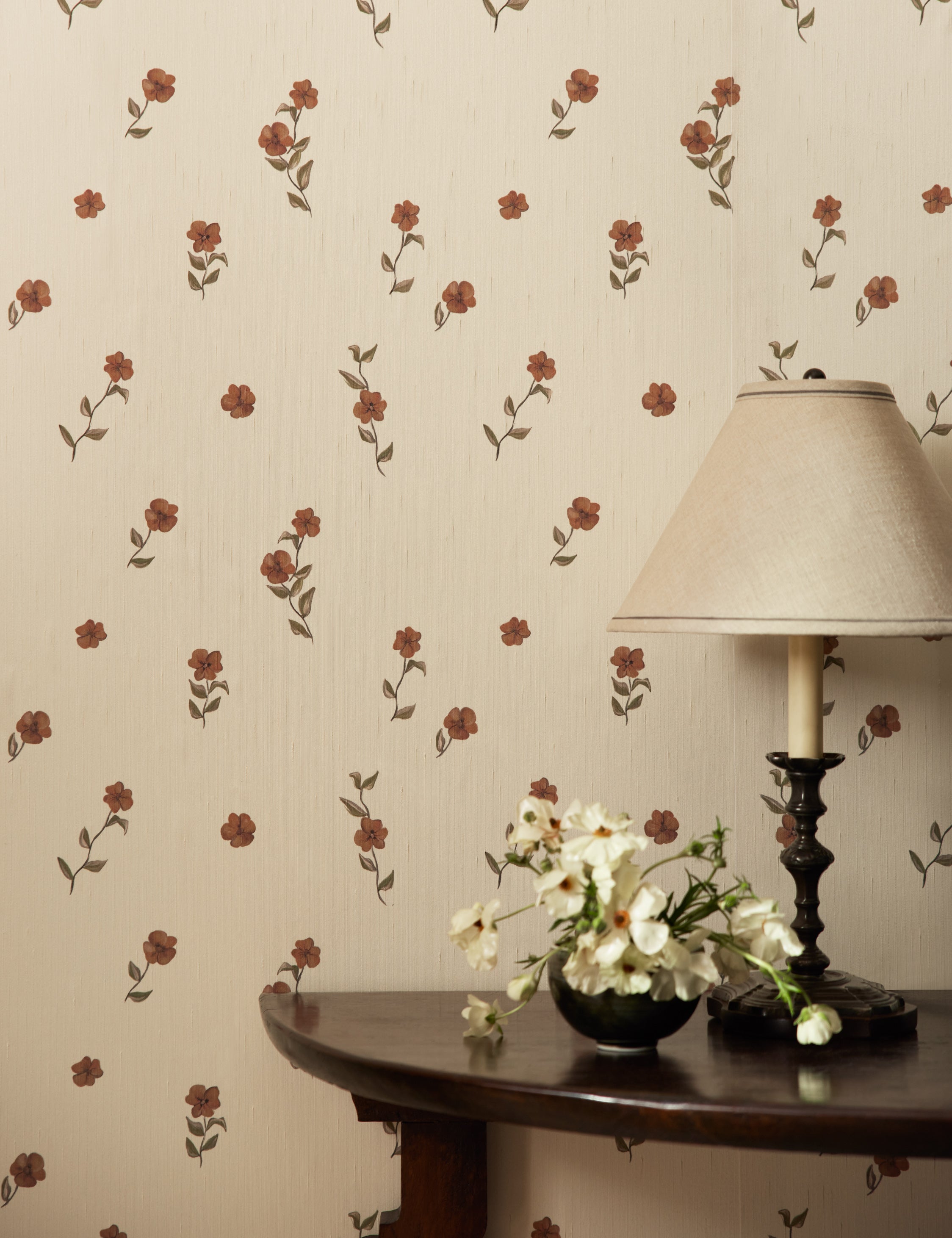 A console table with a table lamp sits in front of a wall covered with the Bamonte Textile String Floral Wallpaper by Jake Arnold