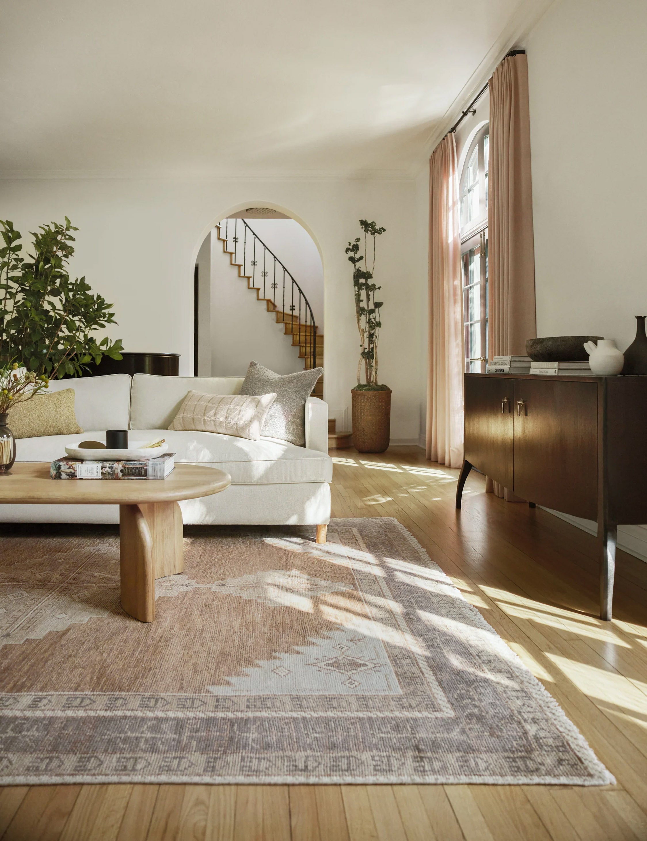 The Kate desert palette geometric floor rug sits in a living room with a white linen sofa, natural wooden coffee table with sculptural legs, and a wooden anabella console table.