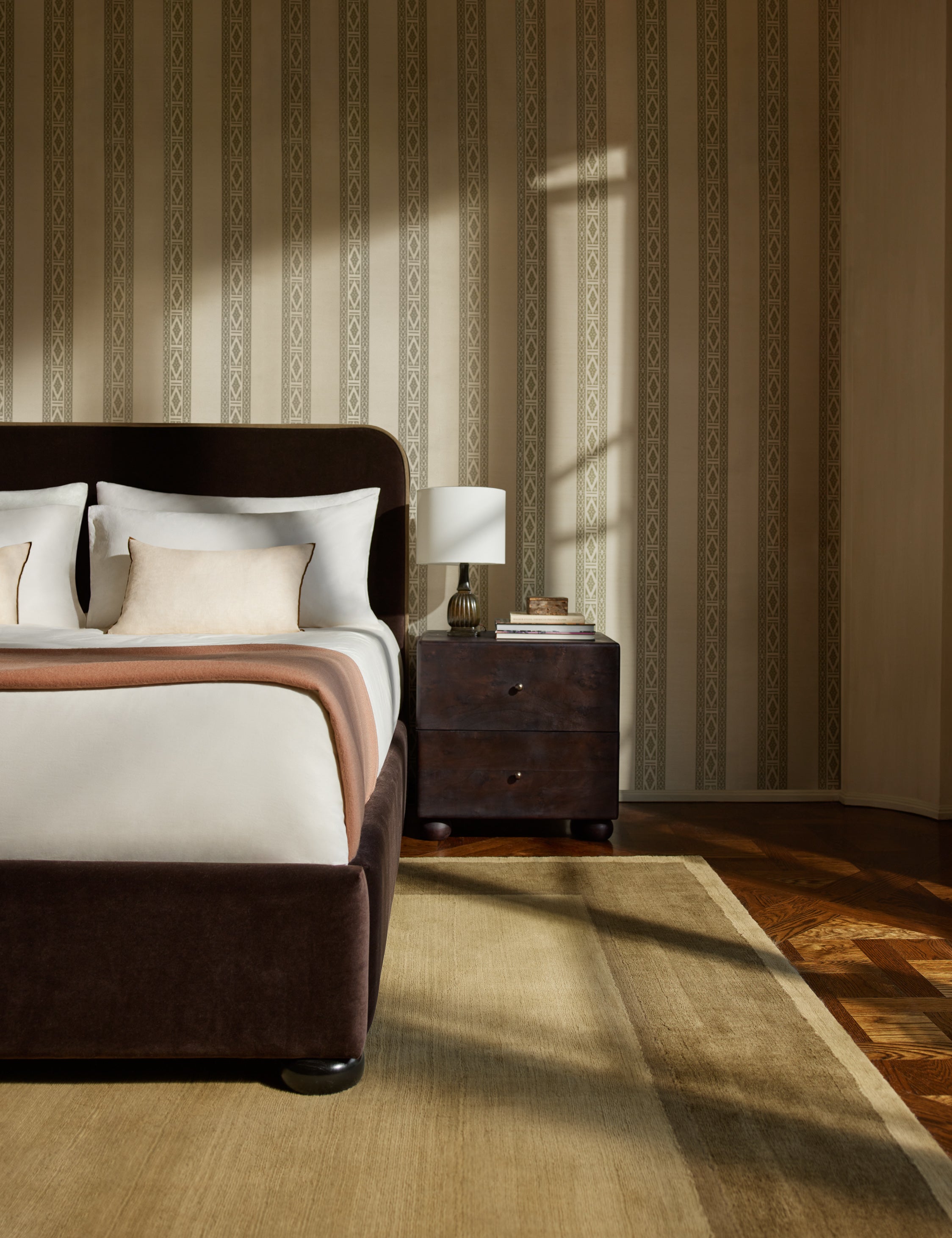 A velvet upholstered platform bed and nightstand sit infront of a wall featuring the Paradiso striped Wallpaper by Jake Arnold