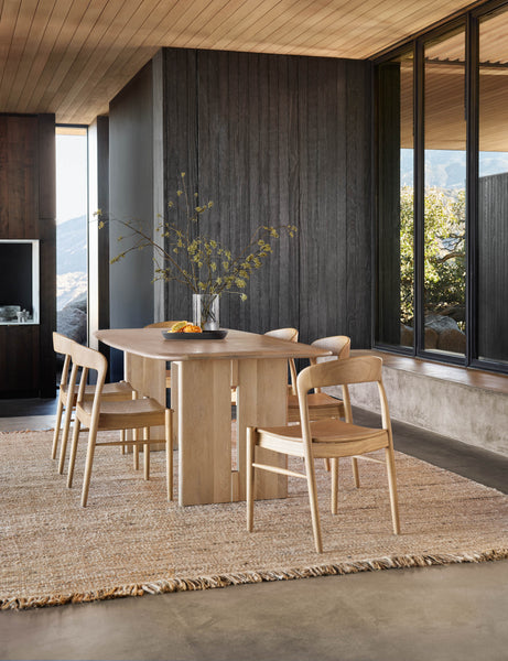 | The Henrik light wood dining table sits atop a jute rug surrounded by six light wood dining chairs in front of a black wood wall