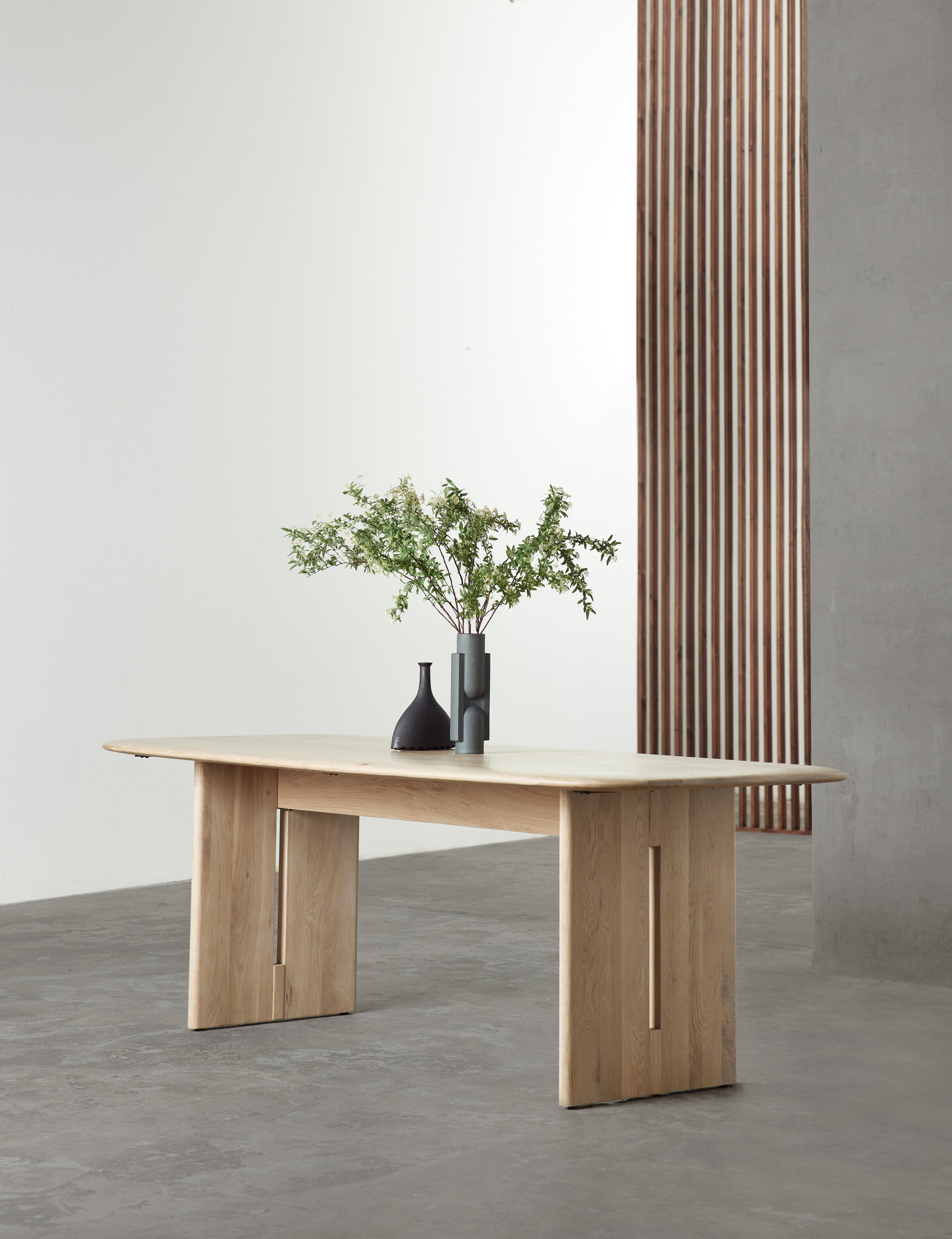 The Henrik light wood dining table sits in a studio atop a gray floor with two sculptural black vases sitting atop it