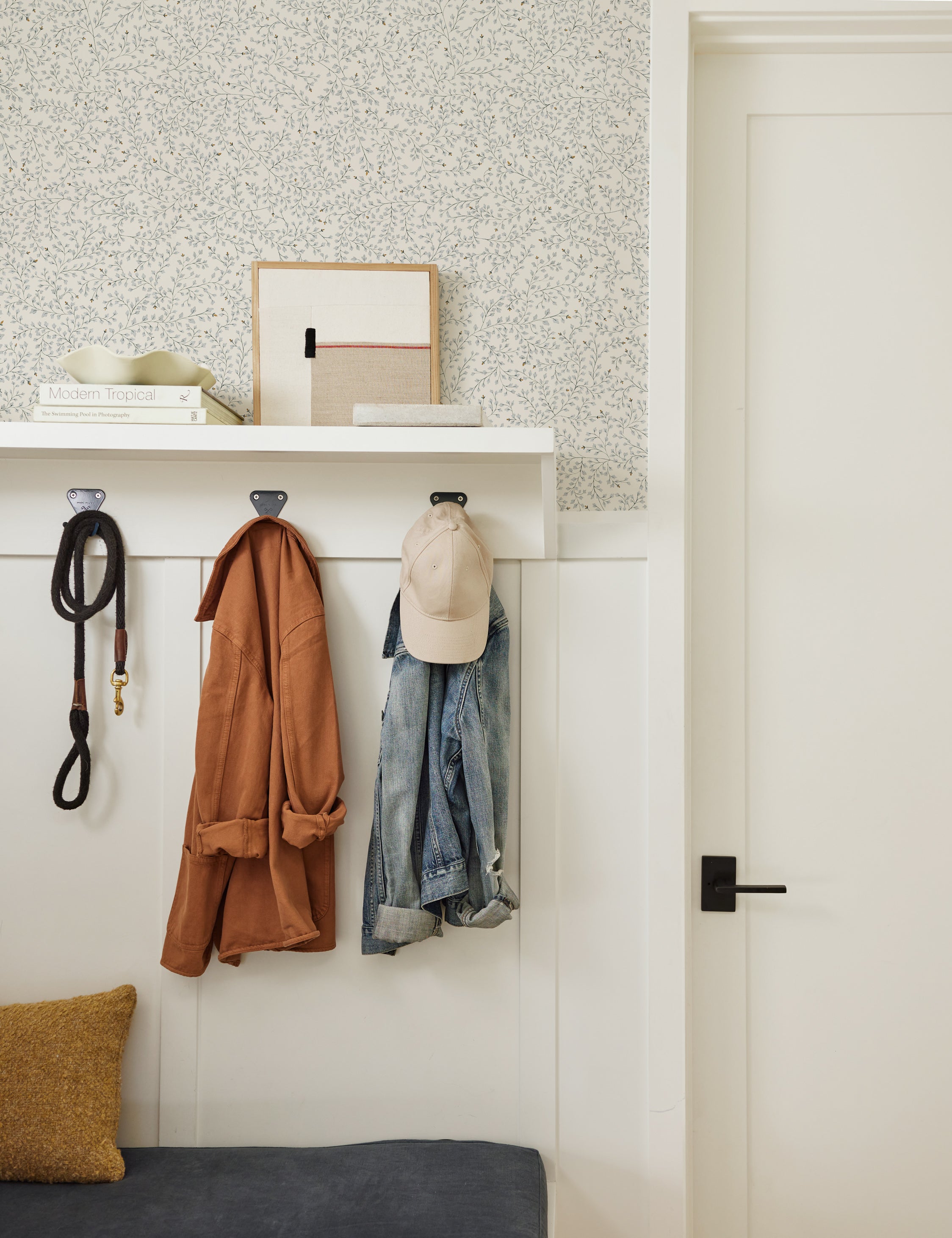 The Dainty Leaves floral Wallpaper by Rylee + Cru is in an entry way with white accented walls, a slate gray cushioned bench with a mustard throw pillow sitting atop it