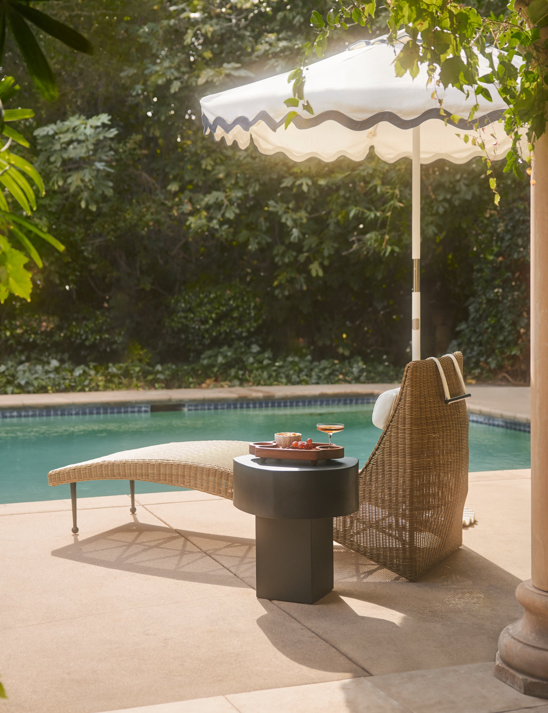 Marisol sculptural wicker outdoor chaise by Sarah Sherman Samuel is styled with an umbrella and black outdoor side table by the poolside.