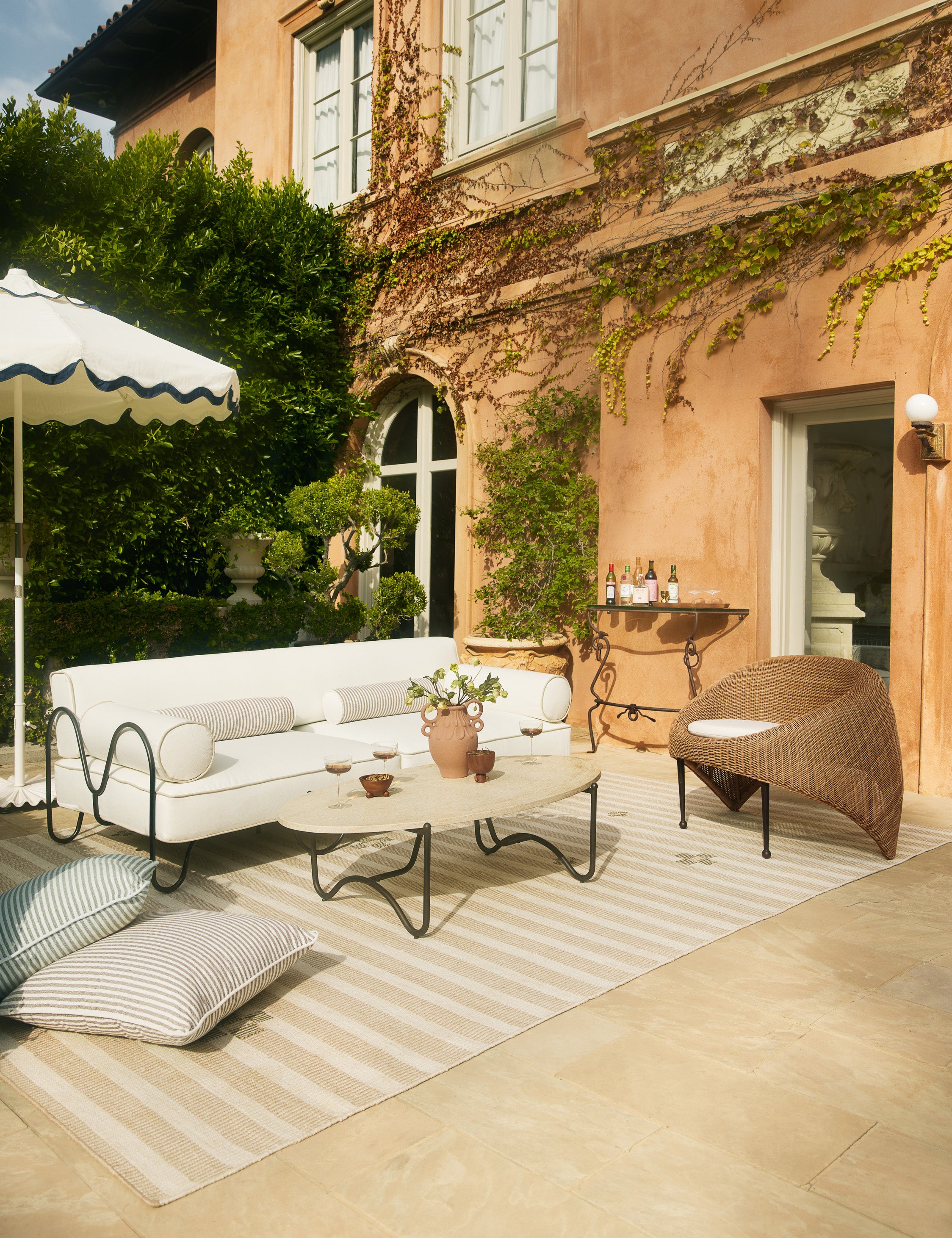 Marisol sculptural wicker outdoor accent chair styled with a modern white outdoor sofa, oval stone top outdoor coffee table and striped outdoor rug on a patio.
