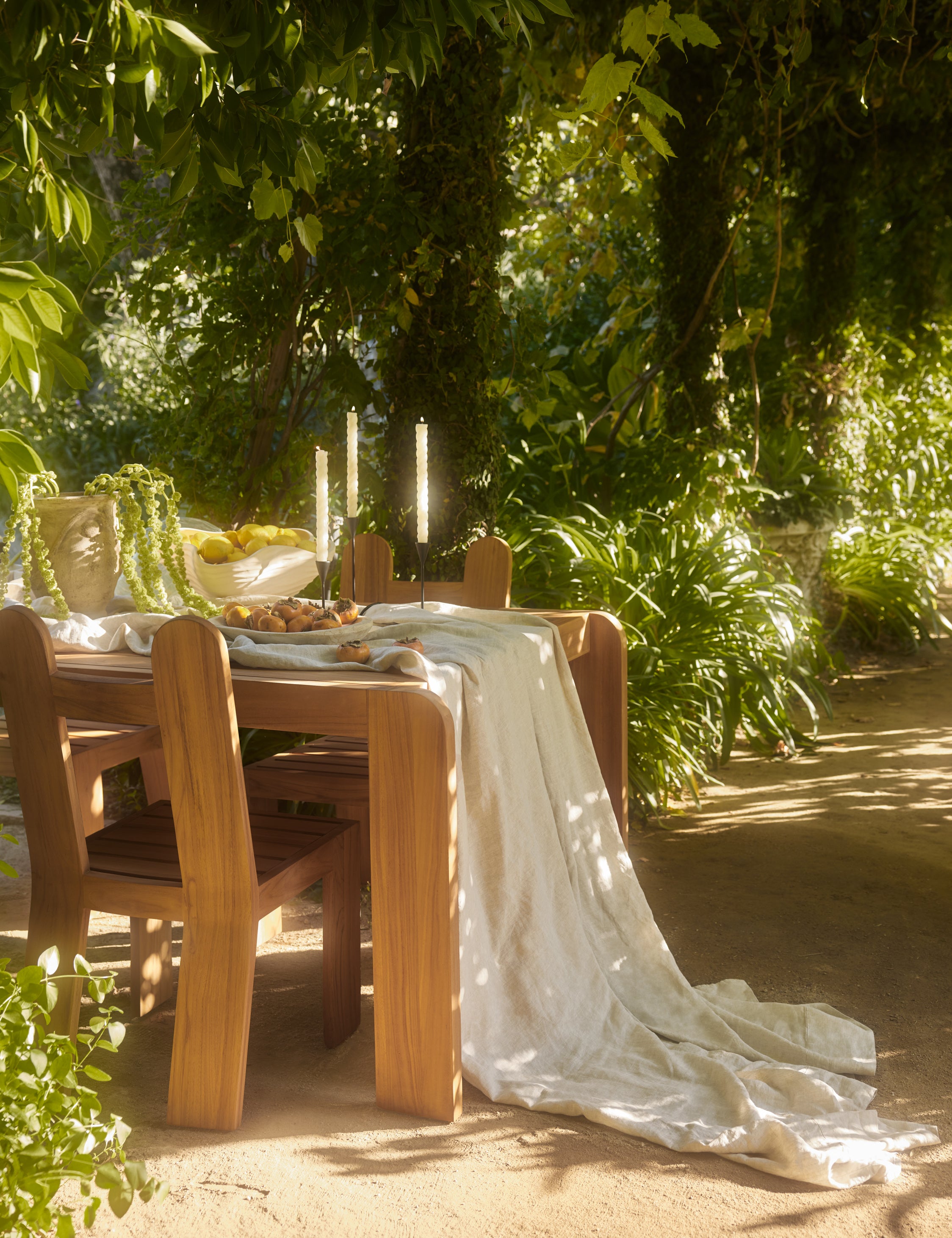 Abbot solid teak sculptural outdoor dining chair by Sarah Sherman Samuel and coordinating Abbot dining table styled with a linen table runner and taper candles.