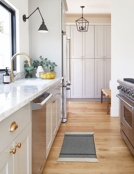 #size::1-9--x-2-10- #size::2--x-5- | The Ines handwoven black-and-white striped mat lays on a wooden floor in a kitchen with white cabinetry and marble countertops 