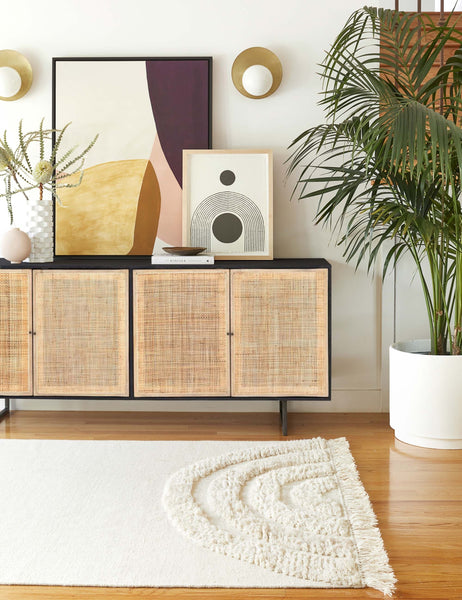 #color::black-and-natural | The Hannah black mango wood sideboard with cane doors and an iron base sits in a room with sculptural wall art sitting atop it and a plush white rug laying in front.