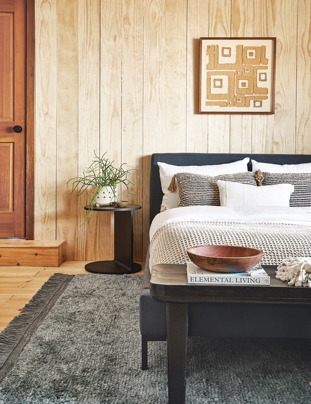 The Illy metal geometric side table sits in a bedroom with a gray linen bed, a gray plush rug, and a light wood paneled wall