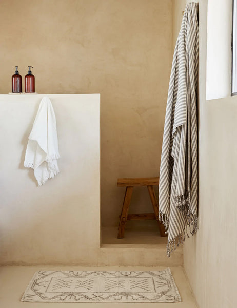 #size::2--x-3- #size::3--x-5- | The Rehya neutral geometric wool patterned rug sits in a bathroom with a wooden stool, a gray and white striped throw, and a white tassel hand towel