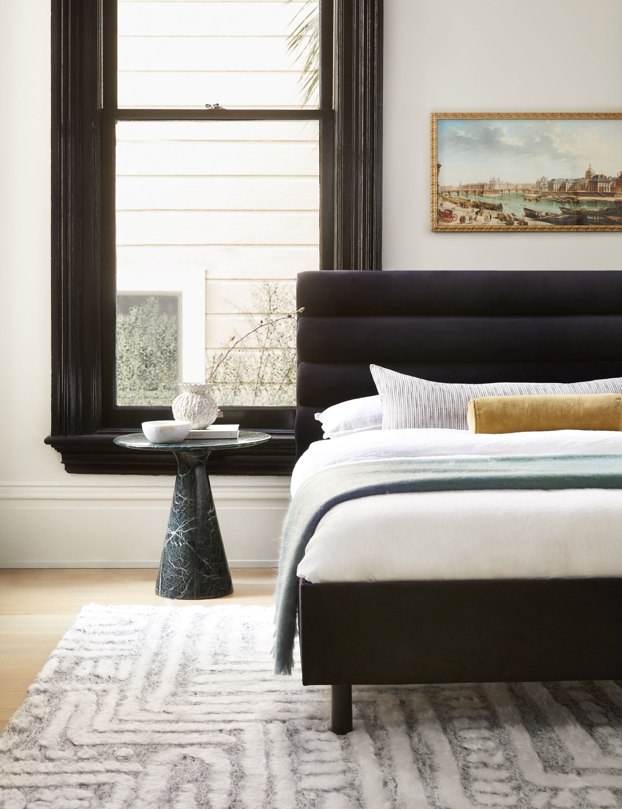 The Peregrine black and white marled striped lumbar pillow lays in a bedroom with a black marble nightstand, a black velvet framed bed, and a landscape painting