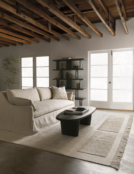 #color::black | The Nera black solid wood sculptural bookcase sits in a living room with an ivory couch and rug, nera black wood coffee table, and a wooden beam vaulted ceiling.