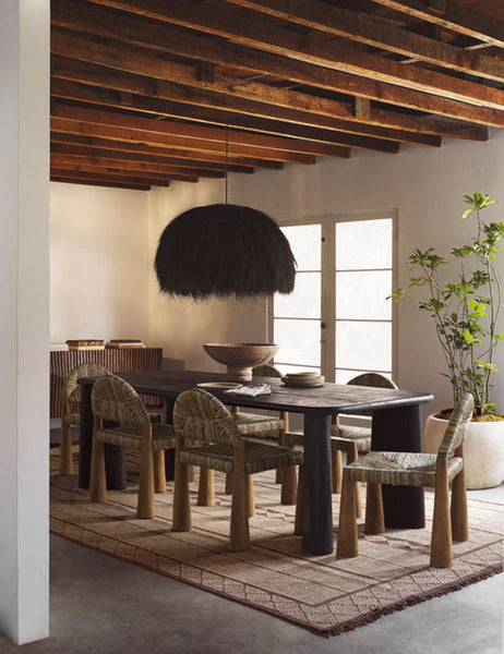 | The Paige black plush pendant light with palm tree fibers sits in a dining room with wooden beamed walls, jute dining chairs, and a brown patterned rug 