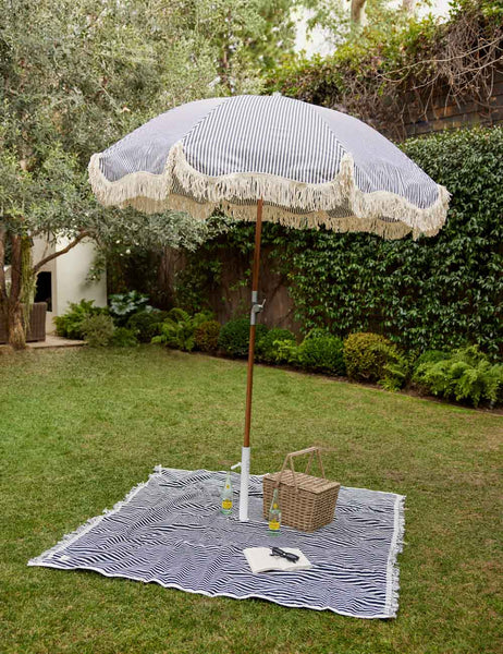 #color::navy-stripe | The Navy and white striped cotton beach blanket by business and pleasure co lays on the grass with a striped umbrella standing atop it