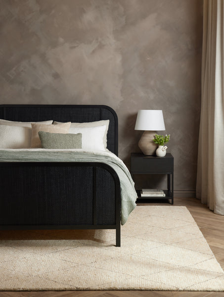 #color::moss #size::13--x-20- | The Manon linen moss green square boucle pillow sits on a black woven framed bed in a bedroom with a plush carpet and brown-toned walls