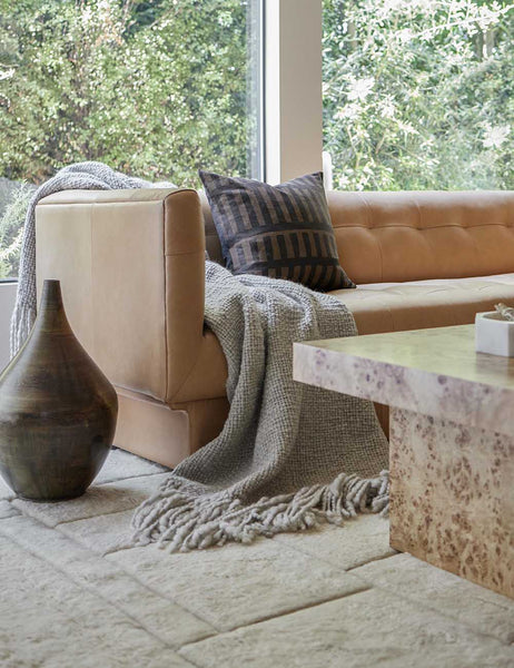 | The Goleta gray chunky wool knit throw blanket with tasseled ends lays on a light brown leather sofa in a living room
