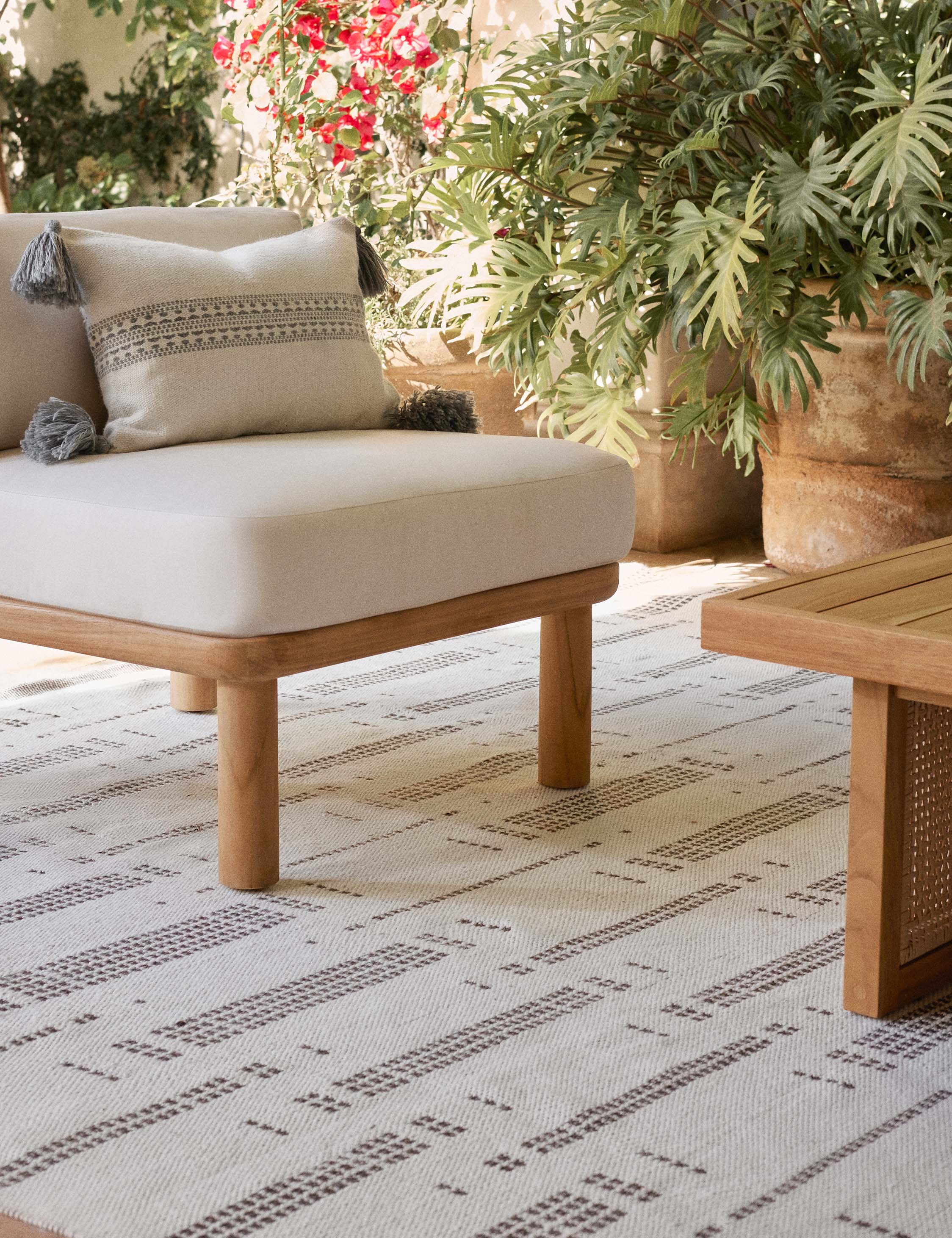 Marchesa natural and agate gray indoor and outdoor lumbar pillow with tasseled corners sits on a sofa in an outdoor space with a natural and brown patterned rug