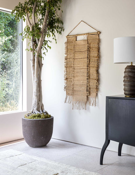 #showall::40271617032291 | The Ukiah woven natural jute and hemp Wall Hanging hangs in a bright room with a ribbed lamp, a black wooden side table, and a plush white rug