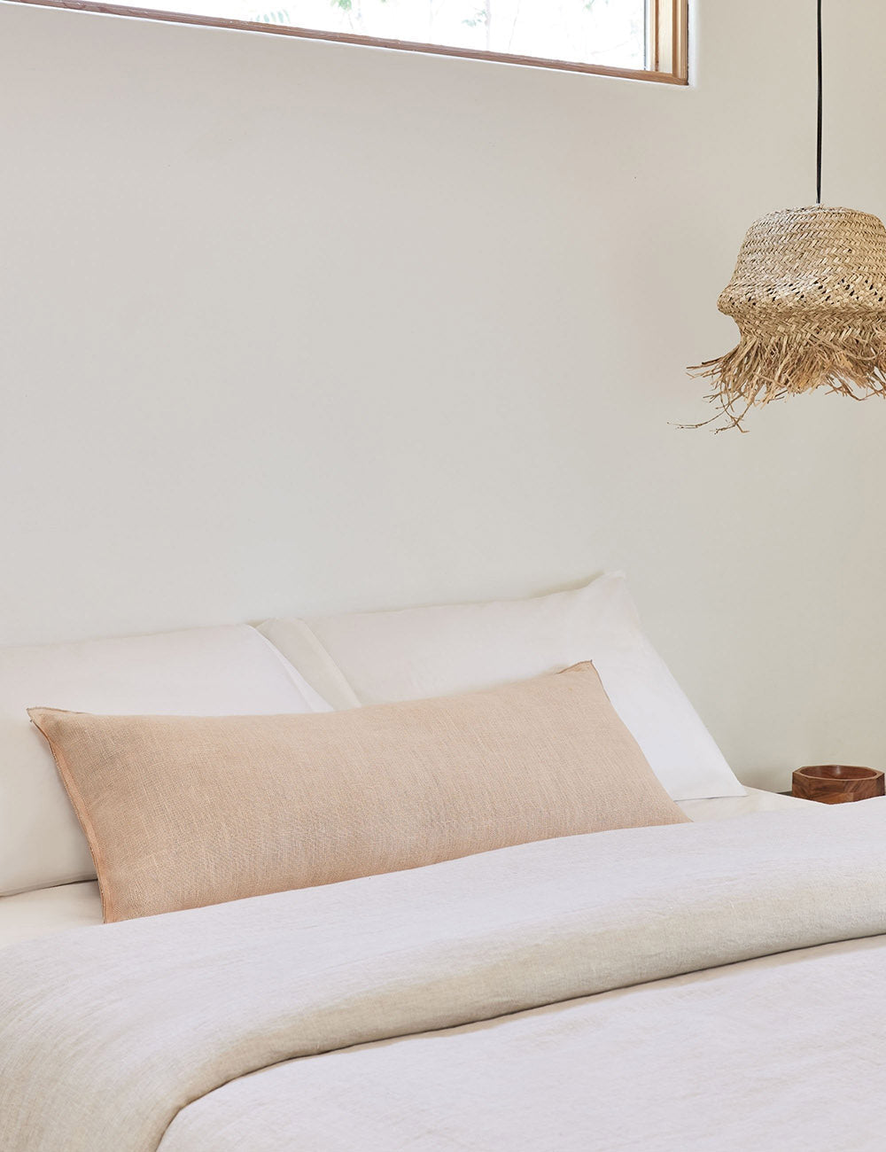 The arlo Blush pink long lumbar pillow lays on a bed with ivory linens with a jute pendant light hanging next to it