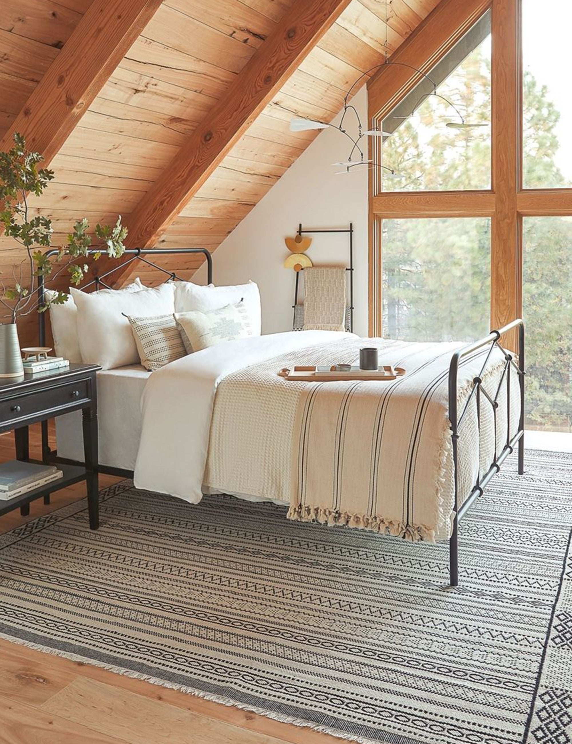 The Monte viso natural and black striped bed cover with fringed ends by House No. 23 sits at the end of a metal framed bed in a bedroom with a geometric rug and sloped wooden ceiling