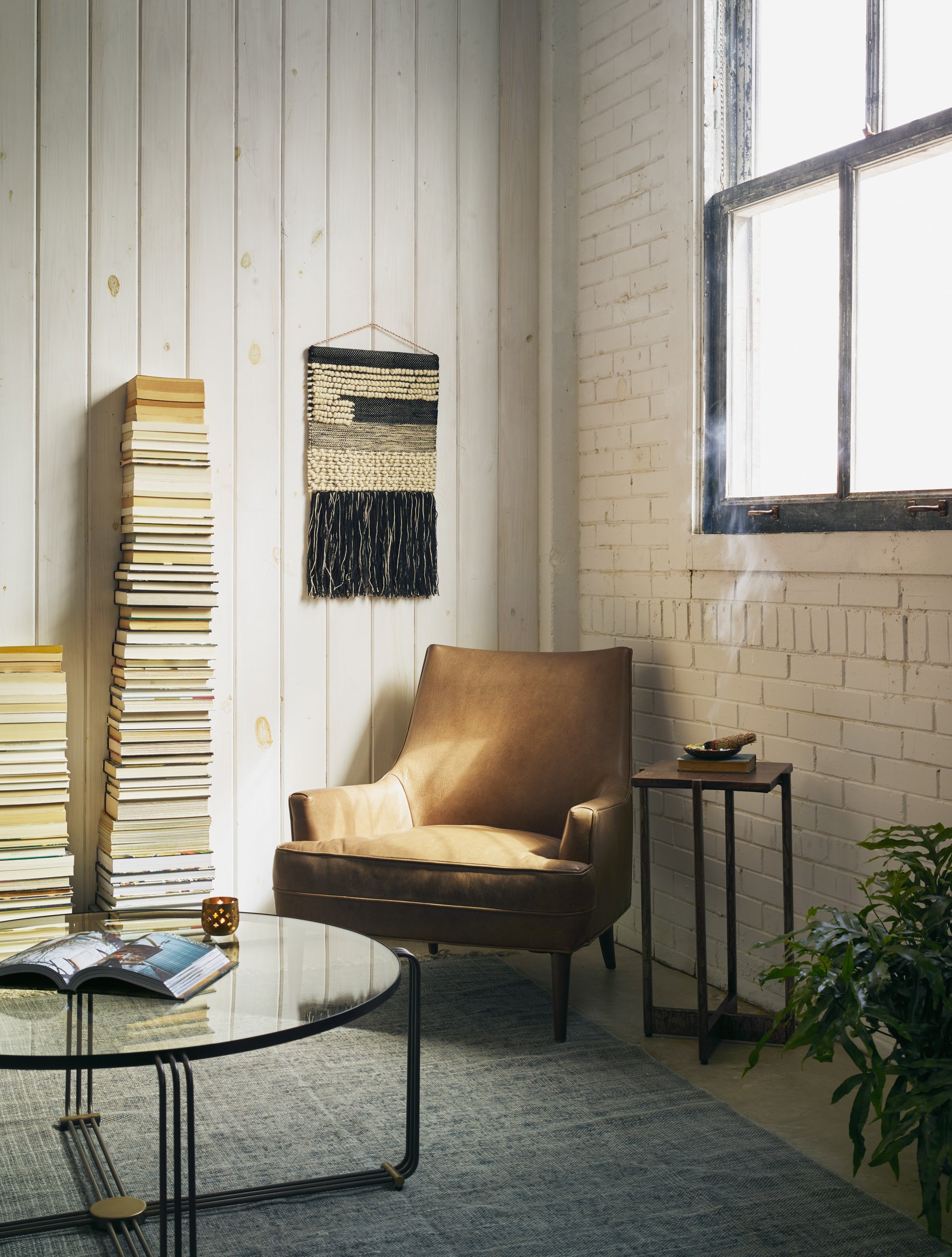 The Ilona Leather Accent Chair sits in the corner of a living room next to a round coffee table and two high stacks of books