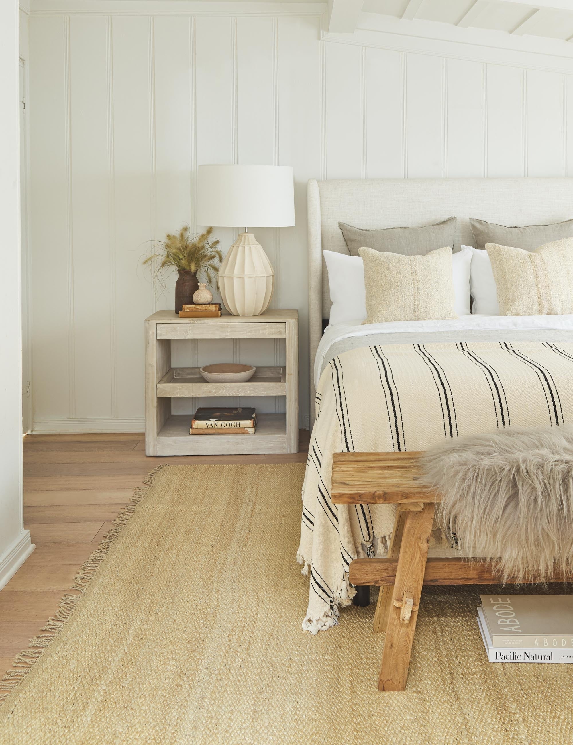 The Monte viso natural and black striped bed cover with fringed ends by House No. 23 sits at the end of a linen adara bed in a bedroom with a jute rug and a white wooden nightstand