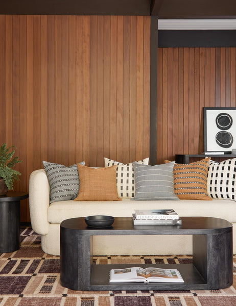 #color::pumice | Bertu pumice gray pillow with a woven dash pattern by Bolé Road Textiles sits on a cream boucle rounded sofa in a retro living room